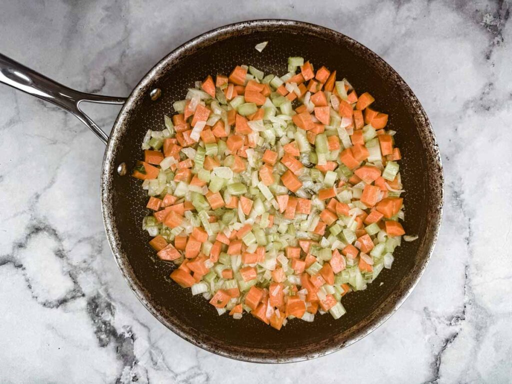 Diced carrots, celery, and onions are sautéing in a frying pan on a marble countertop.
