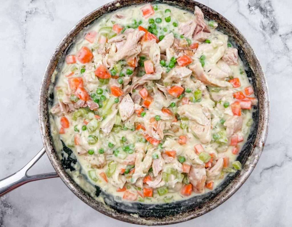 A skillet filled with a creamy mixture of shredded chicken, peas, diced carrots, and celery on a light marble surface.