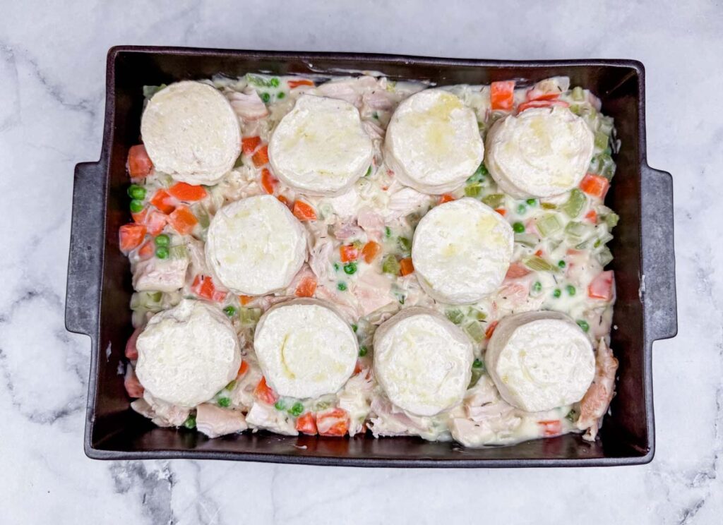 A baking dish filled with a creamy mixture of chicken, peas, carrots, and celery, topped with nine raw biscuit dough rounds, ready to be baked.