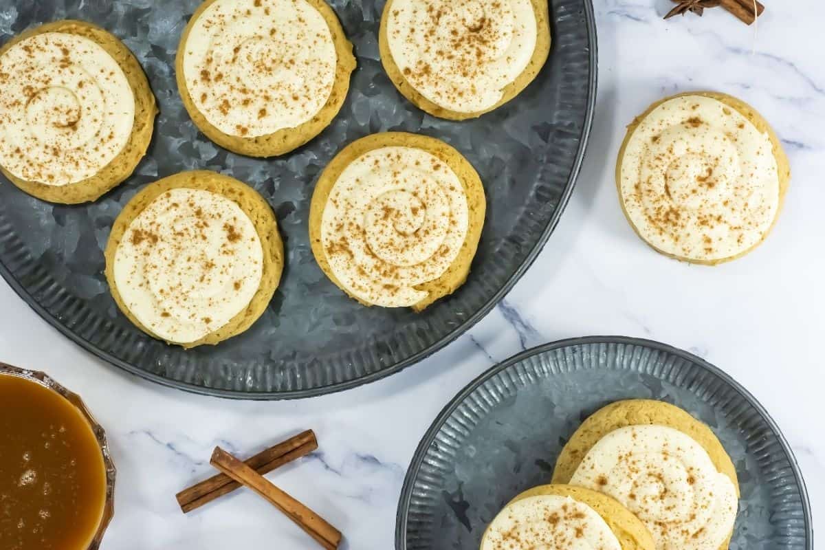 Crumbl Pumpkin Cookies with Caramel are arranged on two metal plates, with cinnamon sticks and a bowl of caramel nearby on a marble surface.