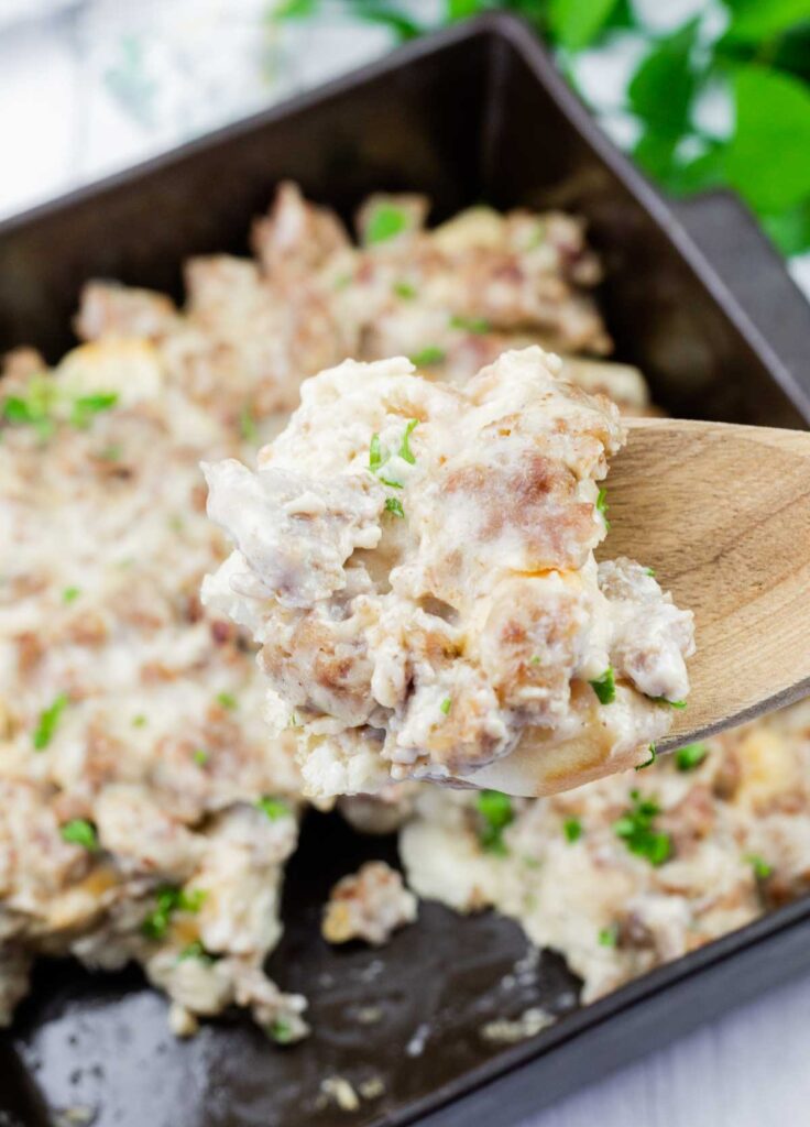 A close-up of a wooden spoon holding a serving of creamy sausage gravy over a baking dish filled with the same mixture, garnished with chopped herbs.