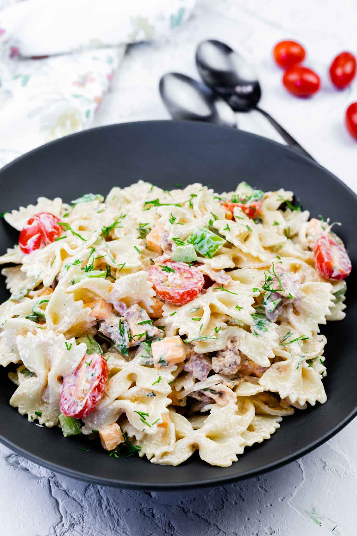 A black bowl filled with bow tie pasta salad mixed with cherry tomatoes, diced vegetables, and a creamy dressing, garnished with chopped herbs.