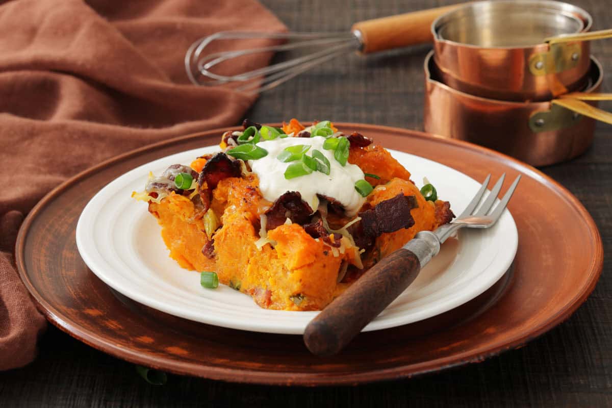 A white plate with loaded sweet potato casserole topped with sour cream, bacon, cheese, and green onions, set on a brown charger plate with a fork and cooking utensils in the background.