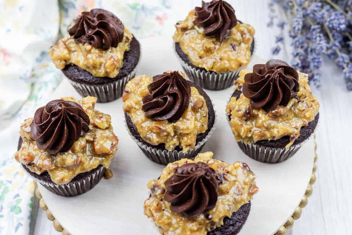 Six German chocolate cupcakes topped with a layer of caramel pecan coconut frosting and a swirl of chocolate frosting, displayed on a round wooden board.