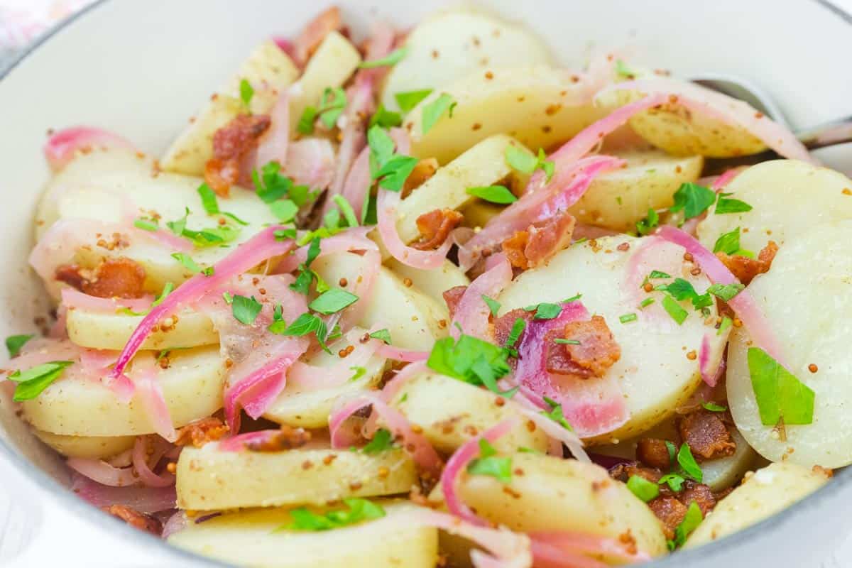 Chef Jenn&rsquo;s Warm German Potato Salad with sliced potatoes with bacon, red onions, and chopped parsley, served in a white bowl.