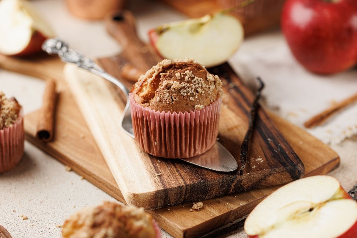 A muffin in a pink paper liner sits on a metal spatula atop a wooden cutting board, surrounded by apple slices and a cinnamon stick.