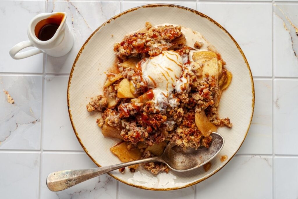 A plate of apple crumble topped with a scoop of vanilla ice cream sits on a white surface, with a spoon and a small pitcher of syrup beside it.