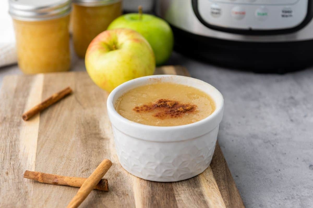 A white ramekin filled with applesauce topped with cinnamon sits on a wooden board next to cinnamon sticks, apples, and an Instant Pot.