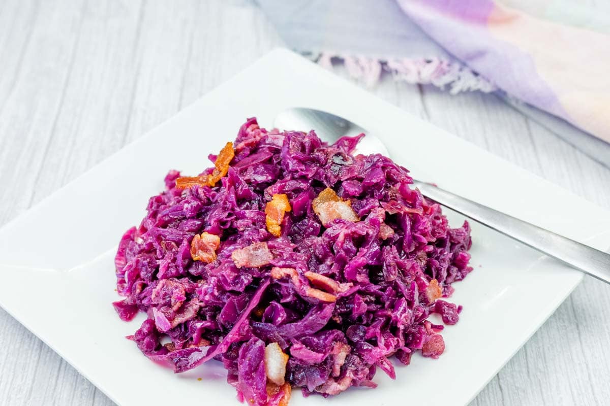 A serving of cooked red cabbage with pieces of bacon and carrots on a white square plate, with a spoon beside the food.