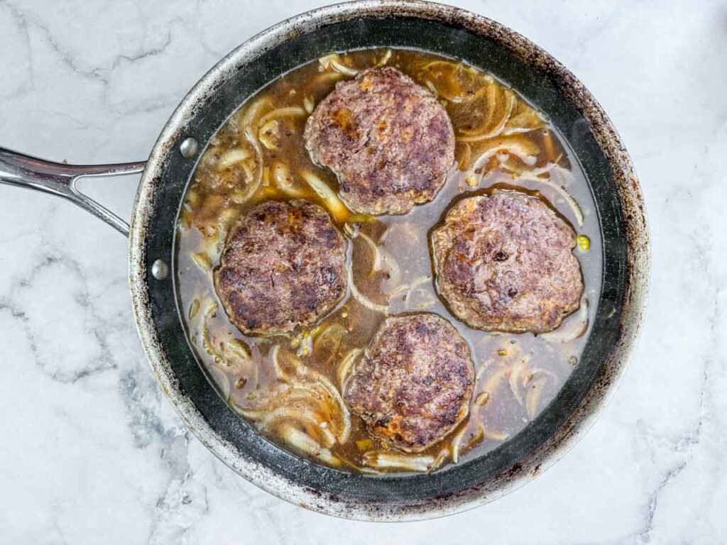 A skillet containing four cooked beef patties simmering in brown gravy with sliced onions.