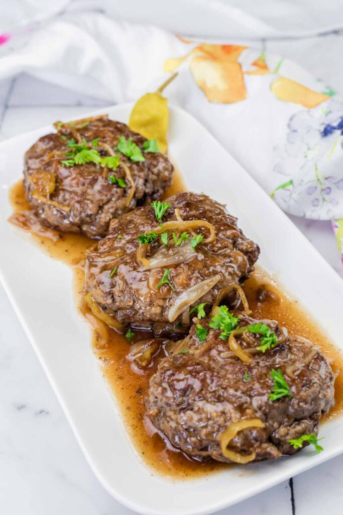 Three hamburger steaks topped with onions and brown gravy, garnished with parsley, served on a white rectangular plate.