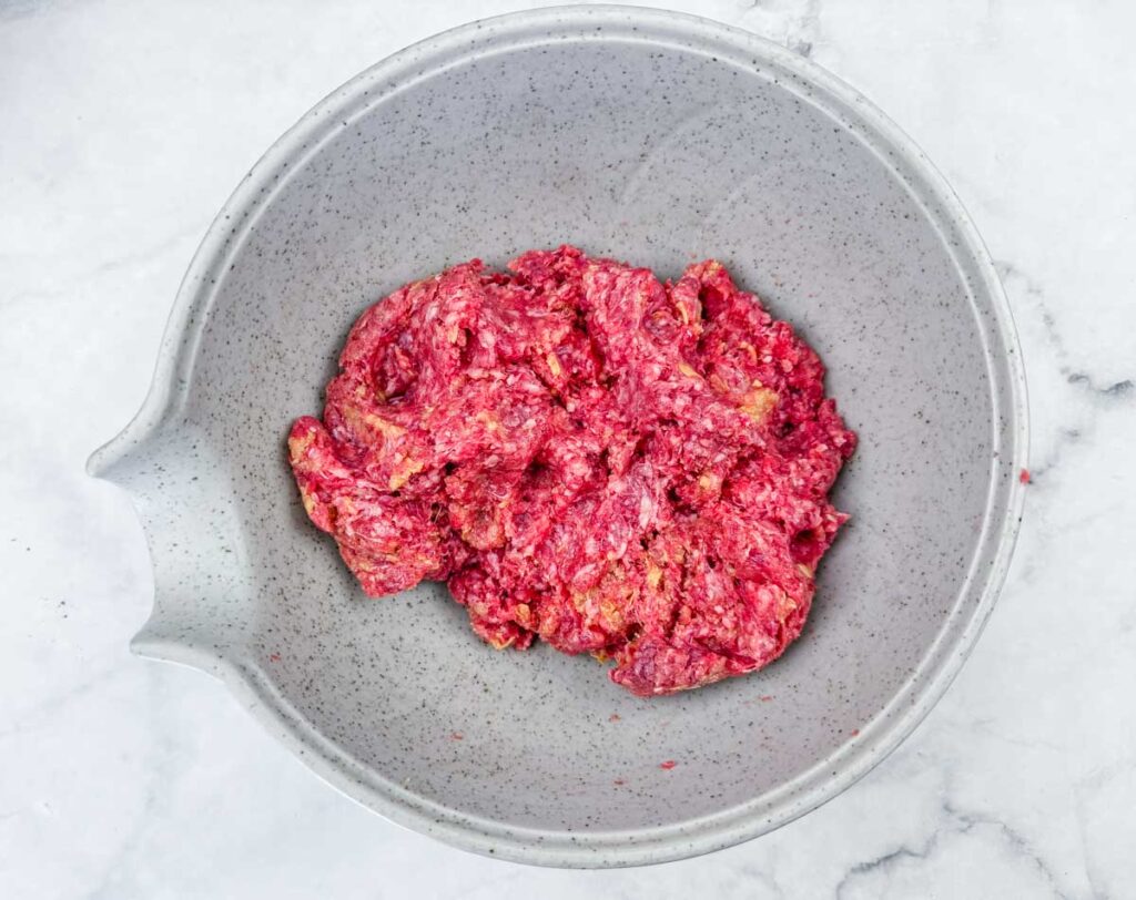 A speckled gray bowl containing a mixture of raw ground meat and seasonings on a light marble surface.
