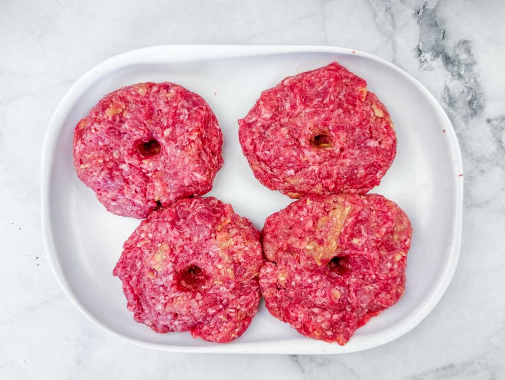 Four raw hamburger patties with holes in the center are arranged on a white oval plate on a marble surface.