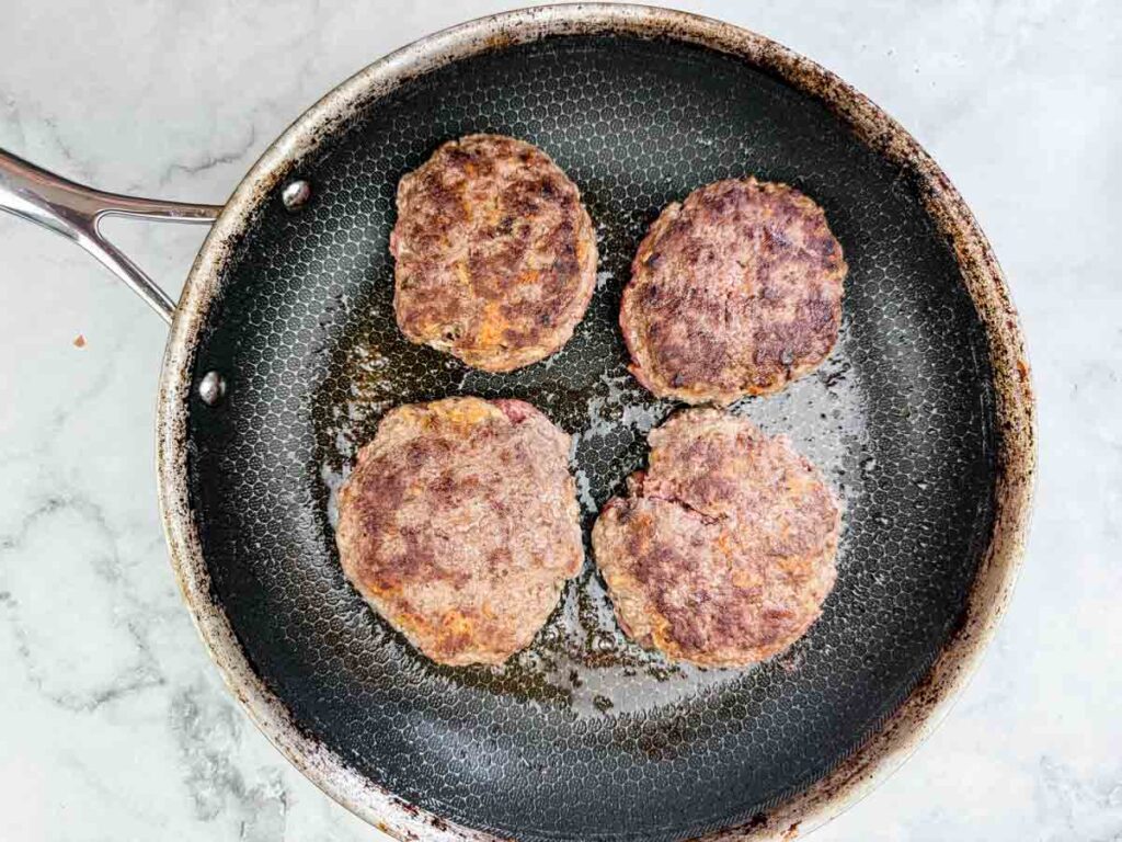 Four beef burger patties cooking in a frying pan on a stovetop, with some oil visible in the pan.