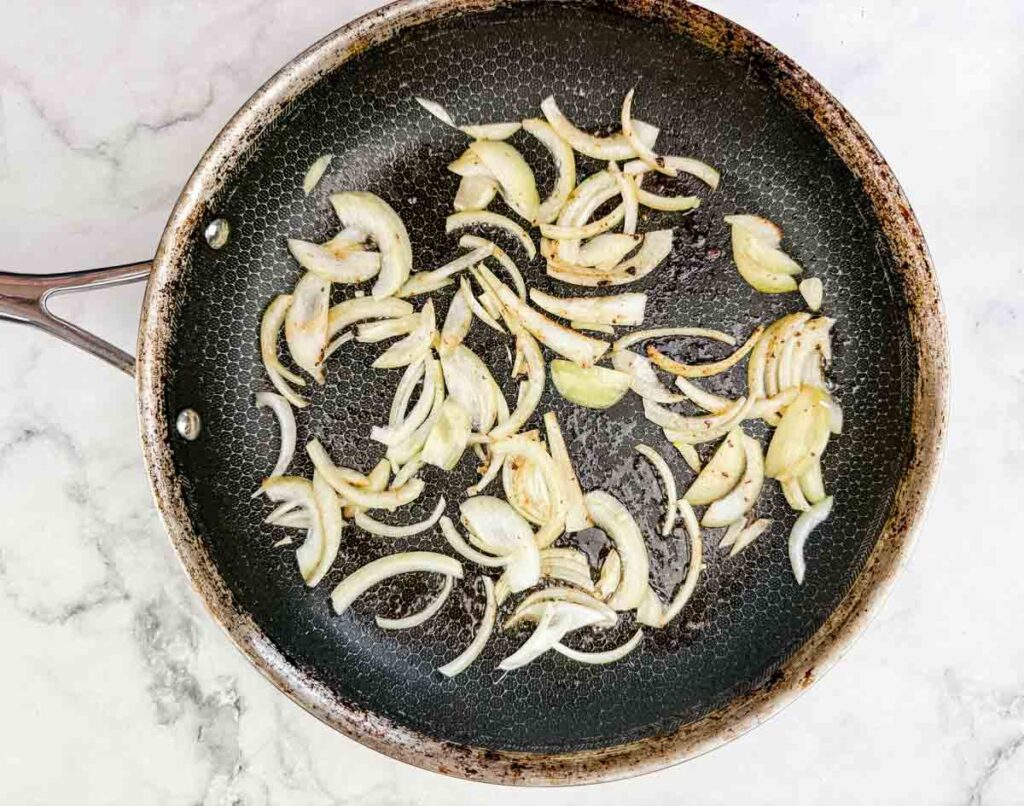 Sliced onions saut&eacute;ing in a frying pan on a white marble surface.