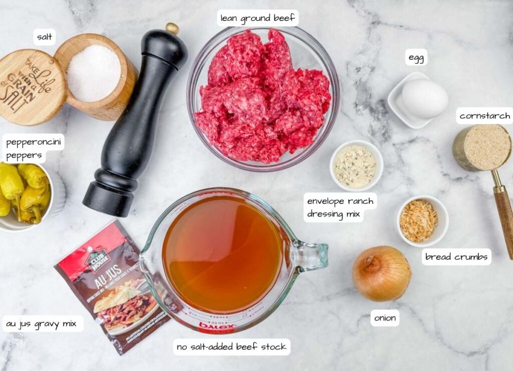 Ingredients for a beef recipe are arranged on a countertop, including ground beef, egg, pepper, salt, onion, bread crumbs, cornstarch, stock, au jus mix, ranch mix, and pepperoncini peppers.