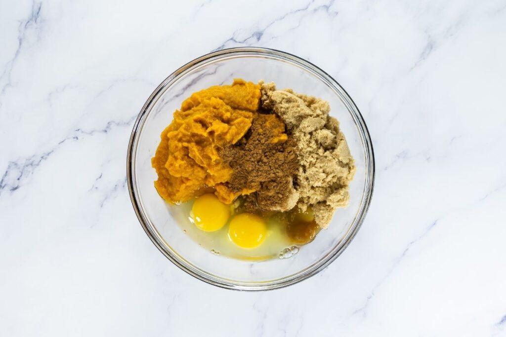 A glass bowl containing pumpkin puree, brown sugar, two raw eggs, and ground cinnamon on a white marble surface.