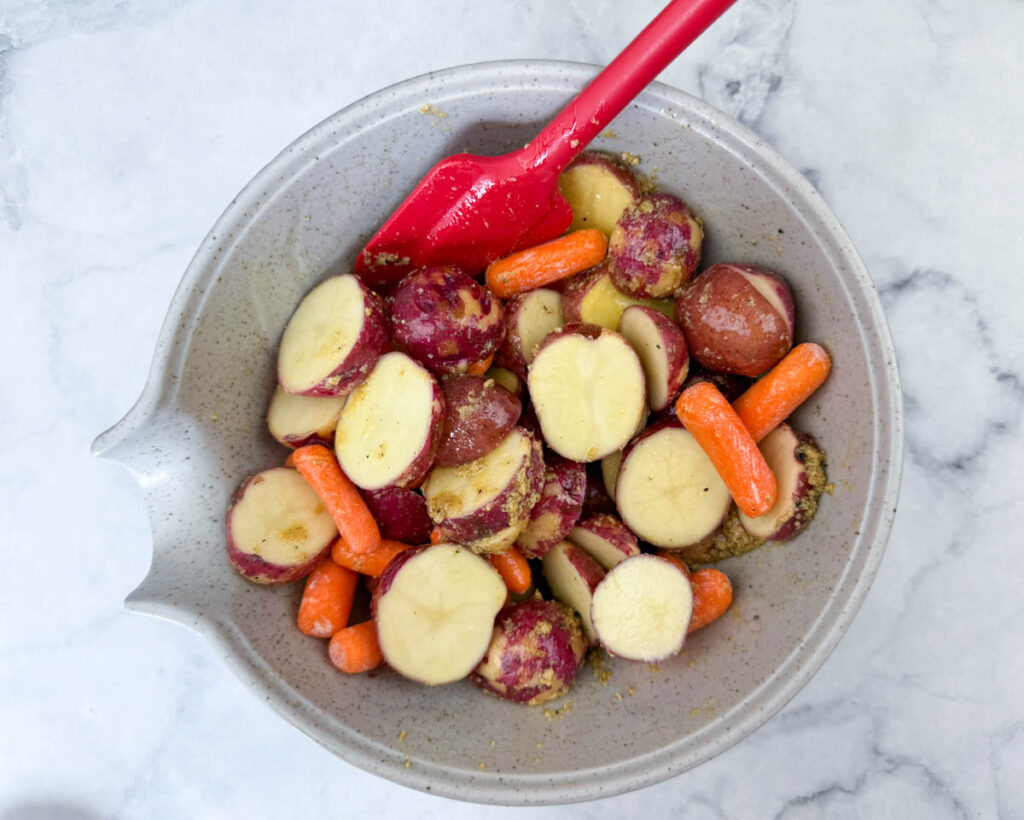 Bowl with sliced red potatoes and baby carrots being mixed with seasoning using a red spatula, on a marble surface.