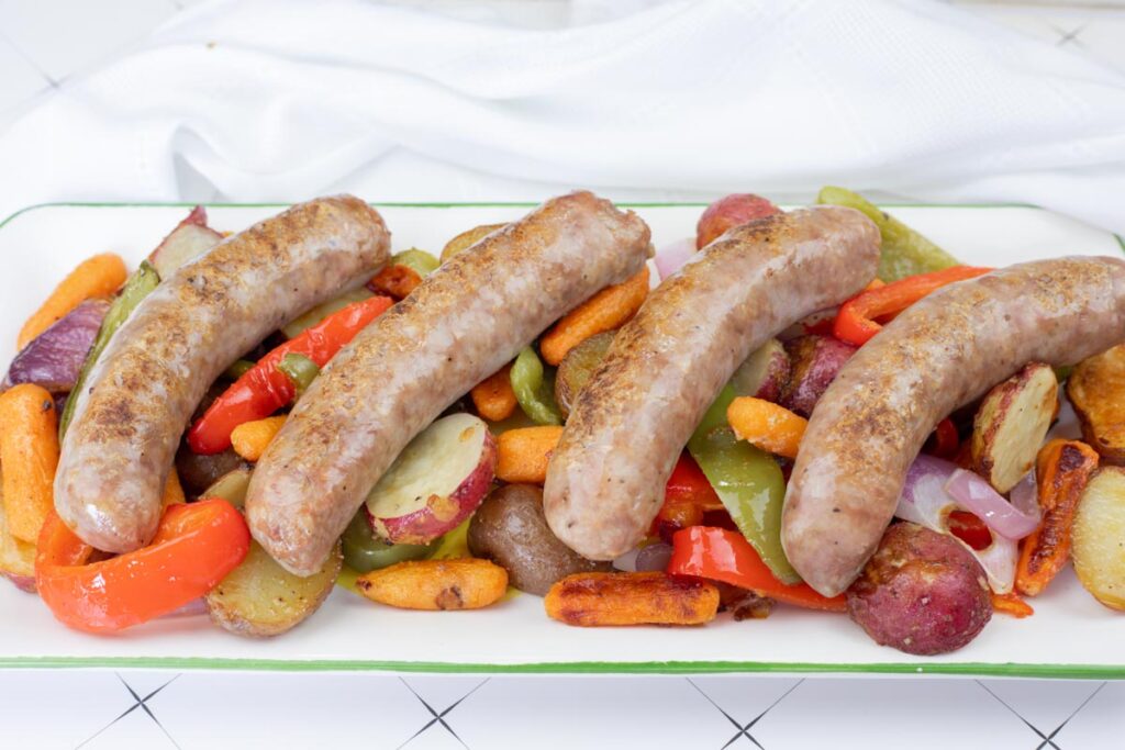 Four cooked sausages are placed on top of roasted vegetables, including carrots, bell peppers, red onions, and sliced potatoes, on a white rectangular platter.
