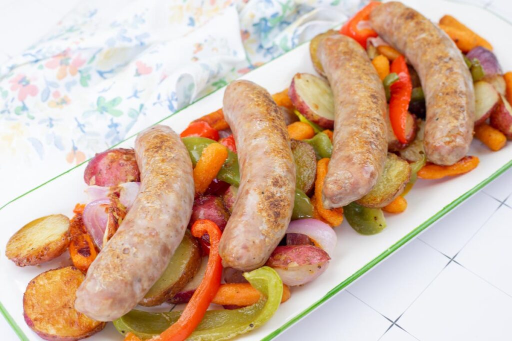 Four cooked sausages served on a platter with roasted potatoes, carrots, onions, red and green bell peppers, placed on a white table beside a floral napkin.