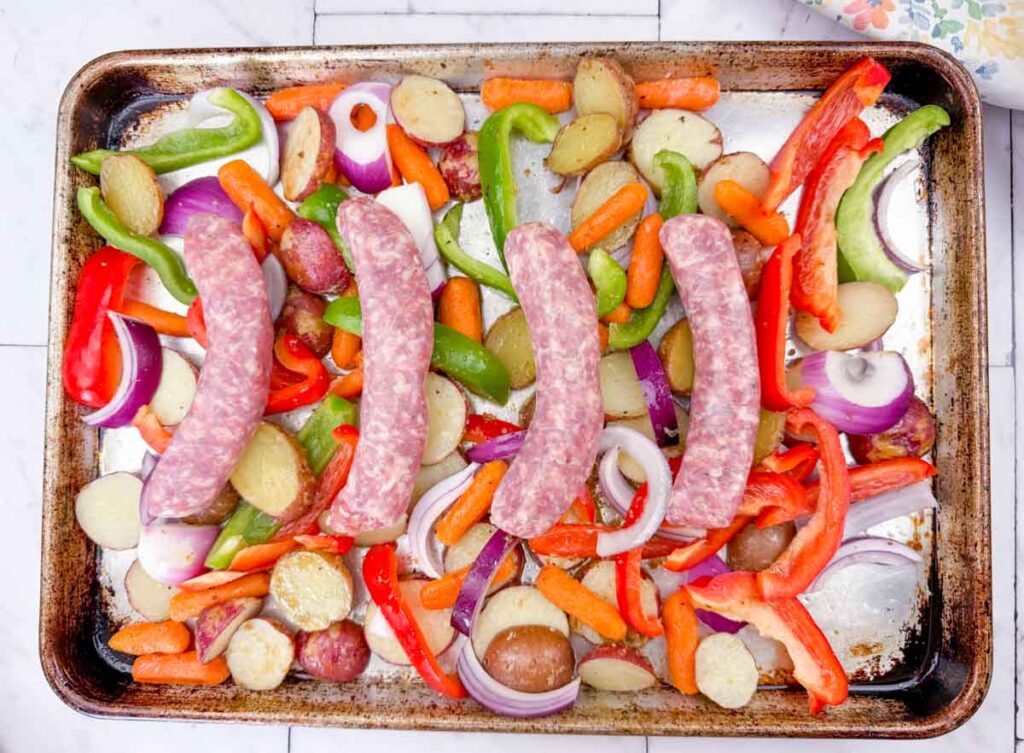 Raw sausages, sliced potatoes, carrots, bell peppers, and red onions arranged on a metal sheet pan, ready for cooking.