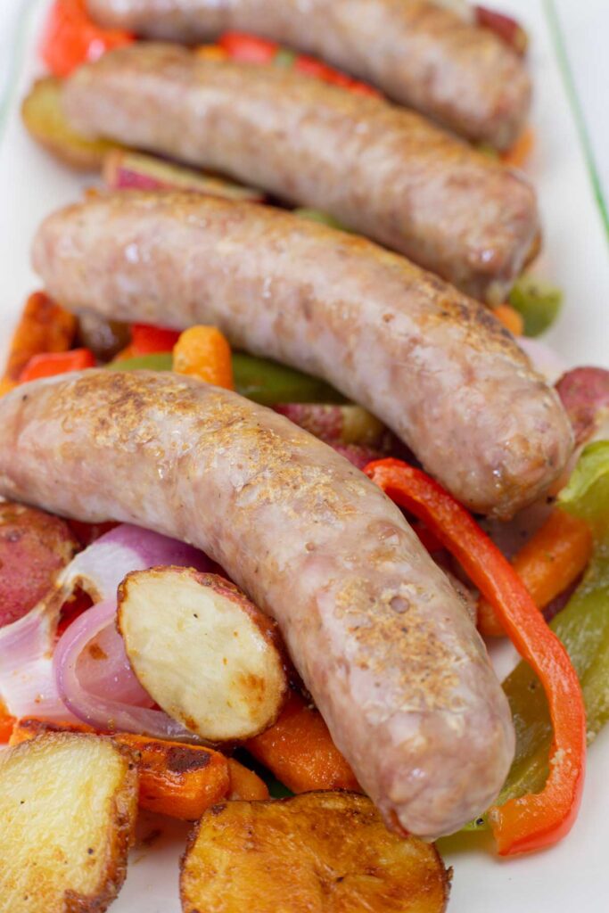 Four cooked sausages served on a bed of roasted potatoes, red onions, carrots, and sliced bell peppers.