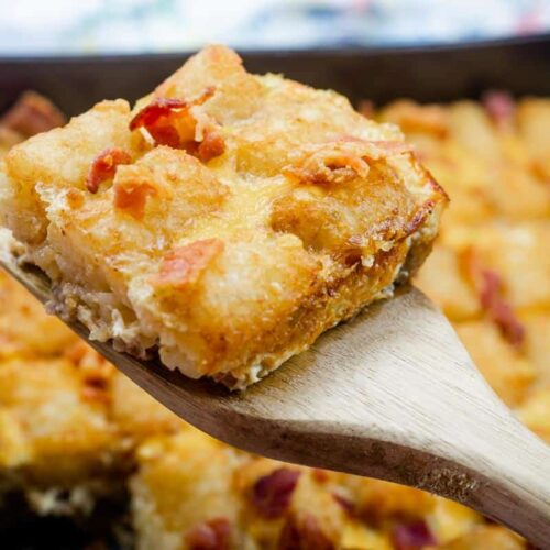 A close-up of a wooden spatula holding a square piece of cheesy breakfast casserole with visible bacon bits, above a baking dish of more casserole.