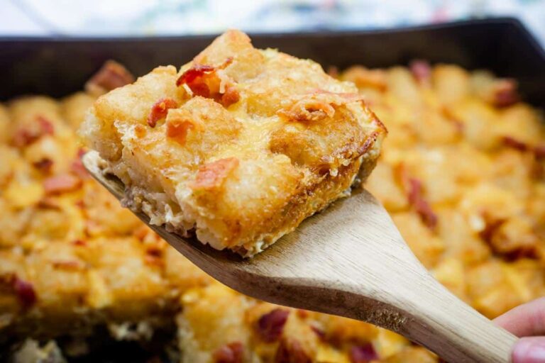 A close-up of a wooden spatula holding a square piece of cheesy breakfast casserole with visible bacon bits, above a baking dish of more casserole.
