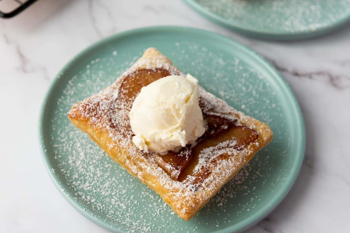 A Puff Pastry Apple Tart topped with a scoop of vanilla ice cream and sprinkled with powdered sugar sits on a light green plate.