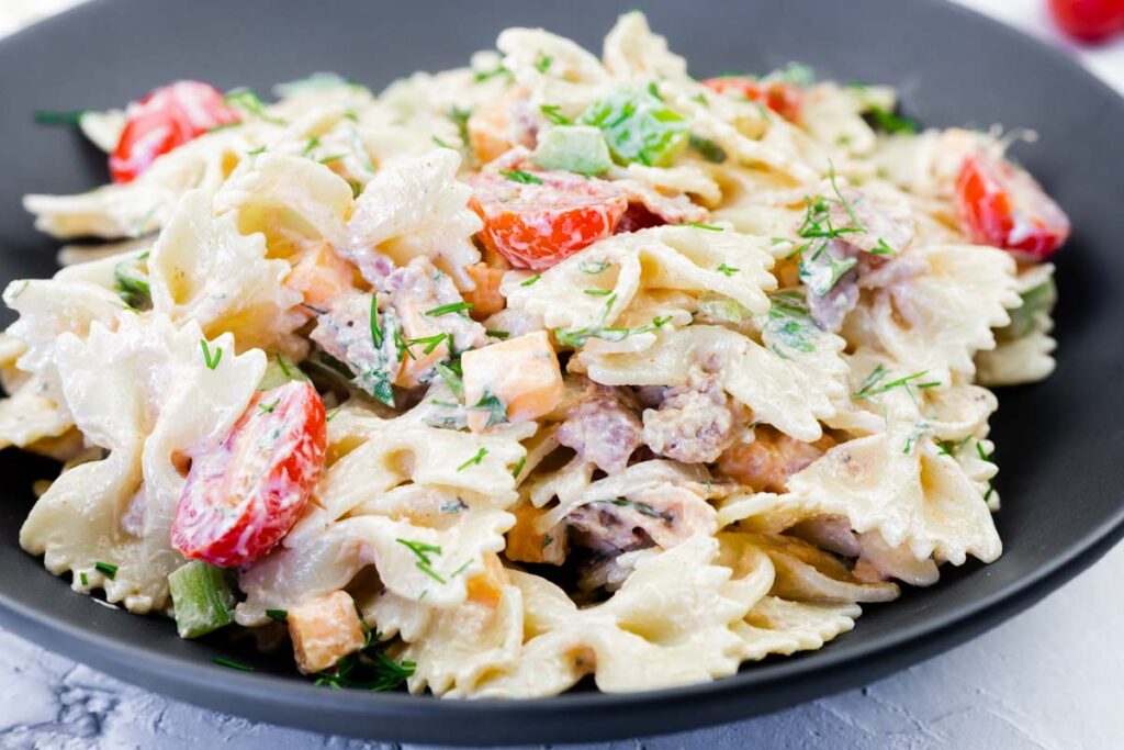 A black plate filled with creamy bow tie pasta salad, mixed with diced vegetables, cherry tomatoes, and herbs.