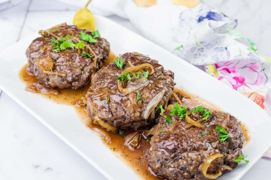 Three Salisbury steak patties with brown onion gravy are served on a white rectangular plate, garnished with chopped parsley.