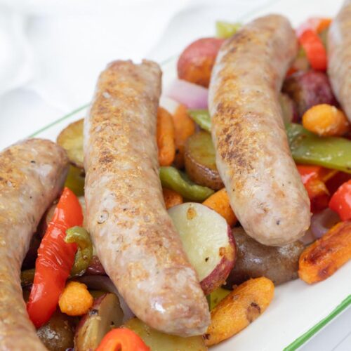 Four cooked sausages served on a platter with roasted potatoes, carrots, onions, green and red bell peppers.
