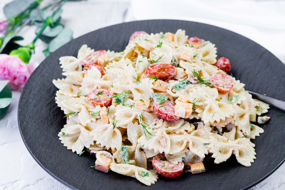 A black plate with farfalle pasta salad mixed with cherry tomatoes, diced vegetables, and a creamy dressing, garnished with herbs.