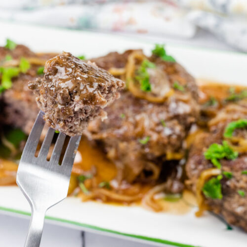 A close-up of a fork holding a bite of cooked ground beef steak with gravy and onions, with more steaks and garnish on a white plate in the background.