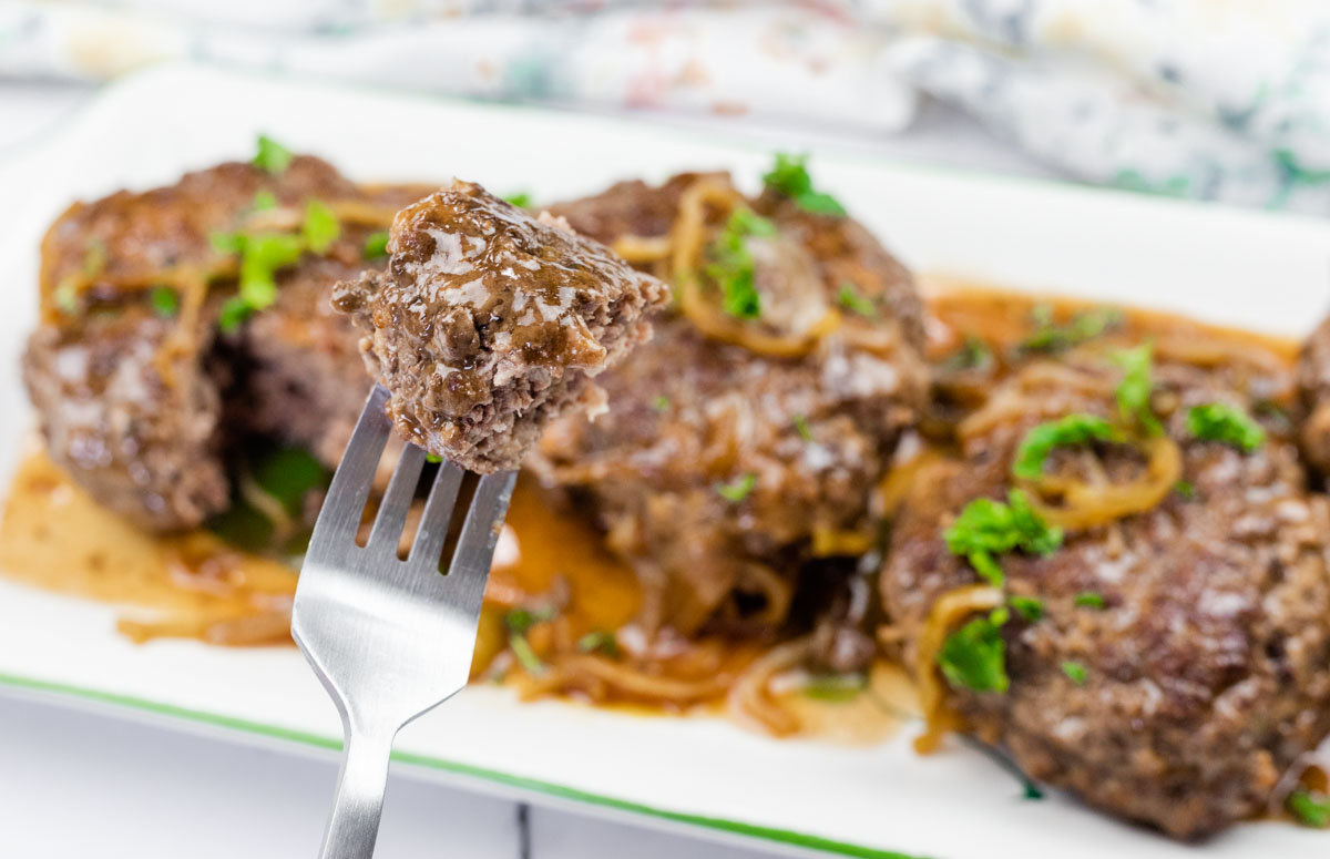 A close-up of a fork holding a bite of cooked ground beef steak with gravy and onions, with more steaks and garnish on a white plate in the background.