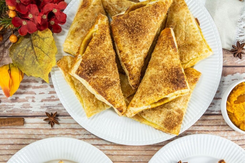 A white plate with several golden-brown, triangular pastry turnovers, surrounded by fall-themed decorations and a small bowl of orange filling.