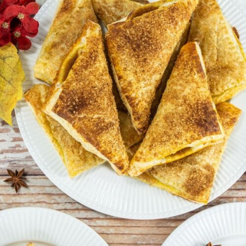 A plate of Pumpkin Turnovers arranged in a stack, with autumn-themed decorations and a small bowl of orange spread nearby.