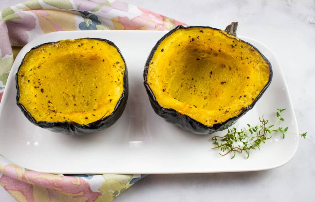Two roasted acorn squash halves on a white rectangular plate with a sprig of fresh herbs, set on a light floral napkin.