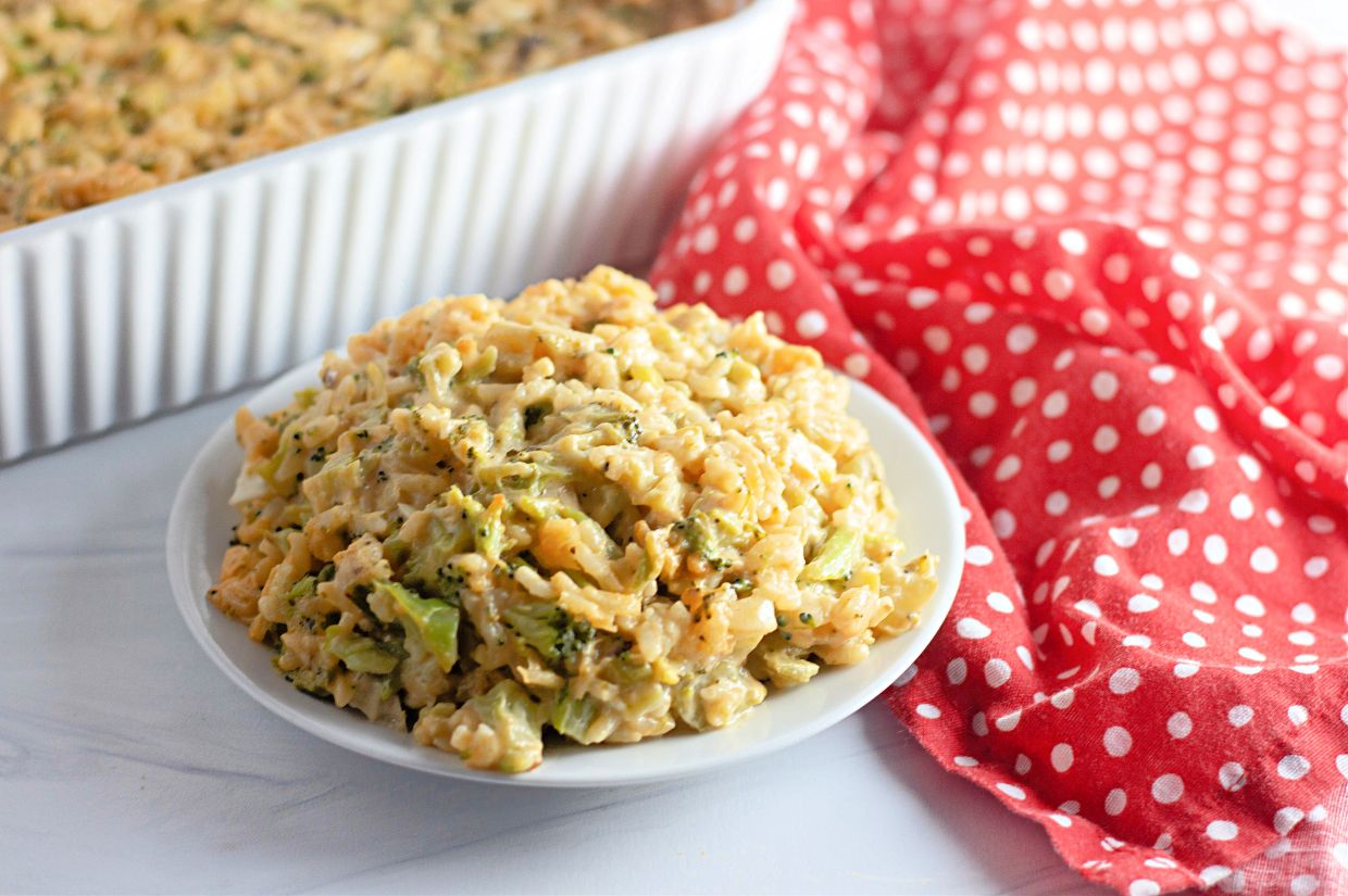 A plate of Easy Cheesy Broccoli Rice Casserole sits on a white surface next to a red and white polka dot cloth, with a baking dish in the background.