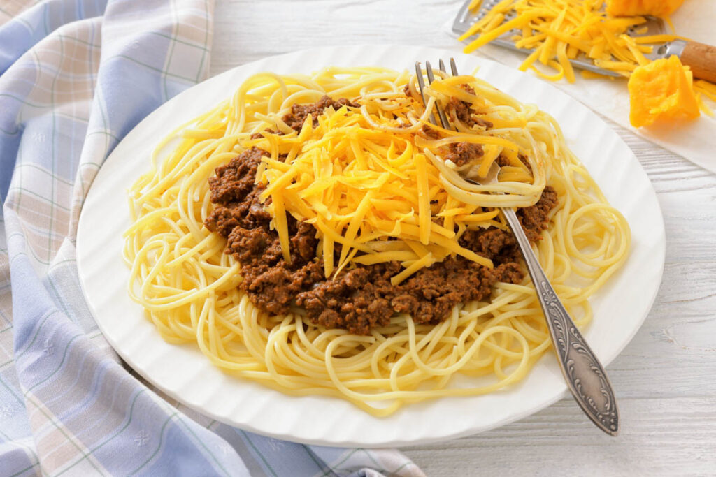 A plate of spaghetti topped with meat sauce and shredded cheddar cheese, with a fork resting on the side; cheese grater and extra cheese in the background.