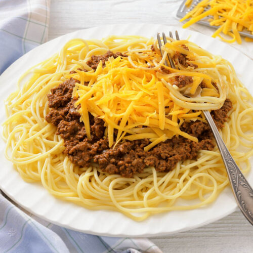 A plate of spaghetti topped with meat sauce and shredded cheddar cheese, with a fork resting on the side; cheese grater and extra cheese in the background.