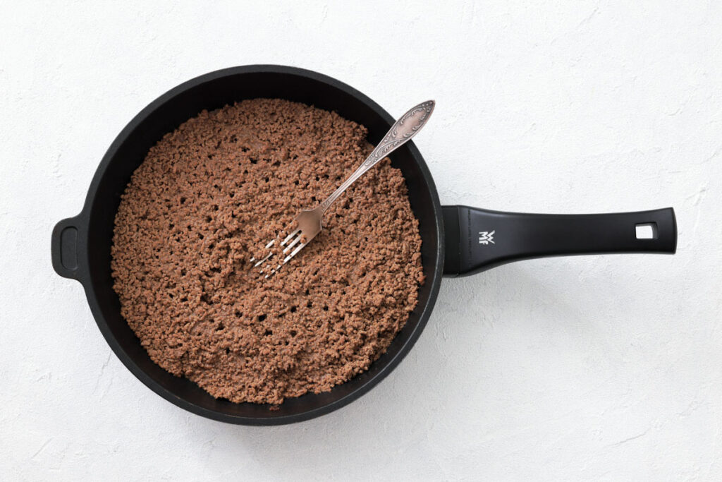 Cooked ground beef in a black frying pan with a metal fork resting on top, placed on a white surface.
