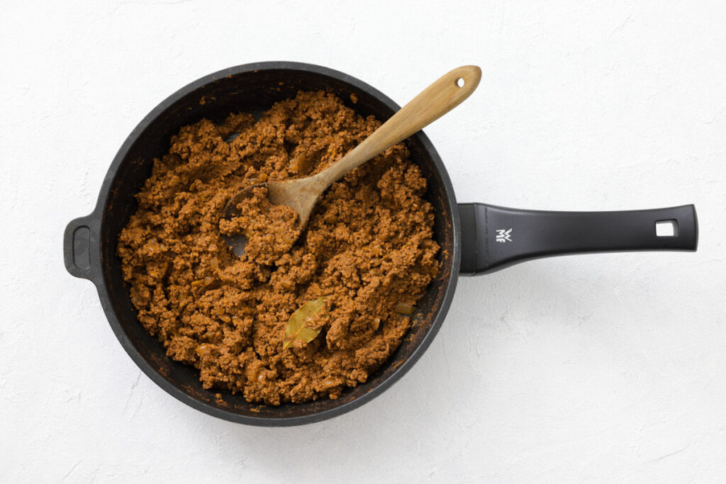 A black pan with cooked ground meat and a bay leaf, stirred with a wooden spoon, placed on a white surface.