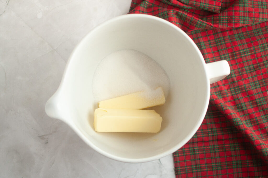 A white mixing bowl with two sticks of butter and granulated sugar inside, placed on a marble surface next to a red plaid cloth.