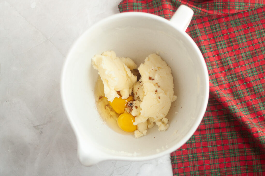 A mixing bowl with creamed butter and sugar, two cracked eggs, and vanilla extract, placed on a marble surface beside a red plaid cloth.