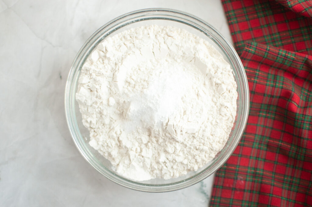 A glass bowl filled with flour and a pile of baking powder or baking soda, placed on a marble surface next to a red plaid cloth.