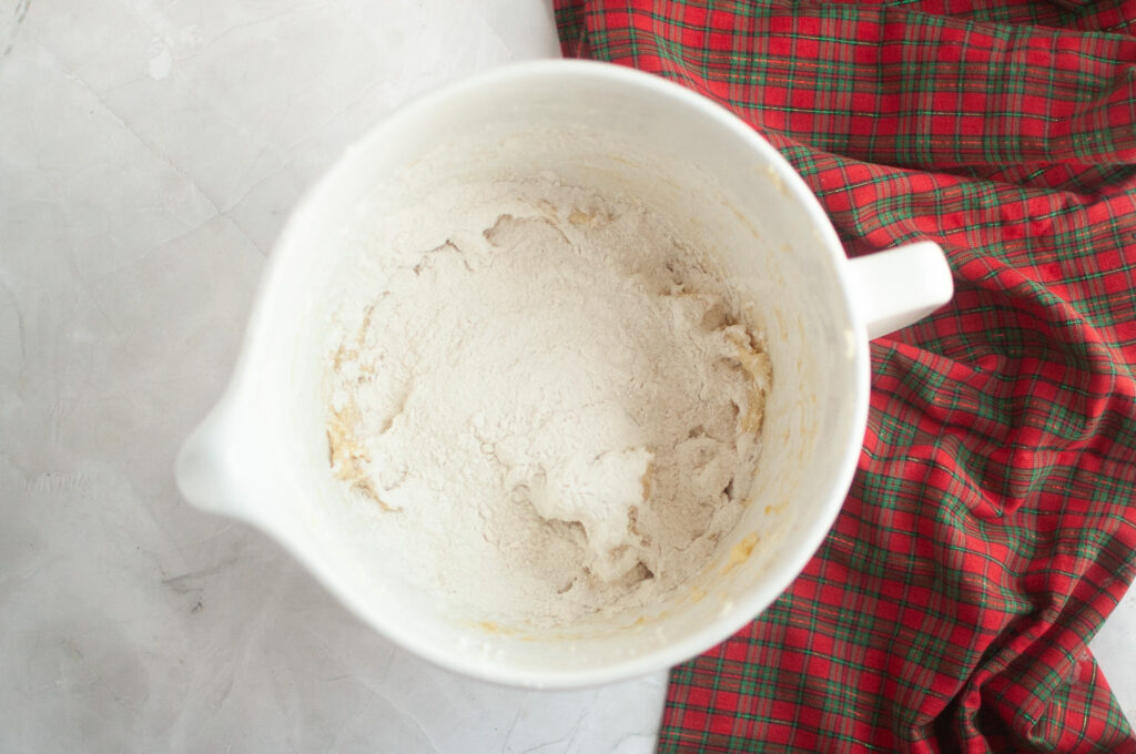 A white mixing bowl with partially mixed dough and flour sits on a marble surface next to a red plaid kitchen towel.