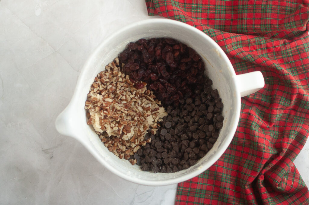 A mixing bowl containing chopped pecans, dried cranberries, and chocolate chips sits on a marble surface next to a red and green plaid cloth.
