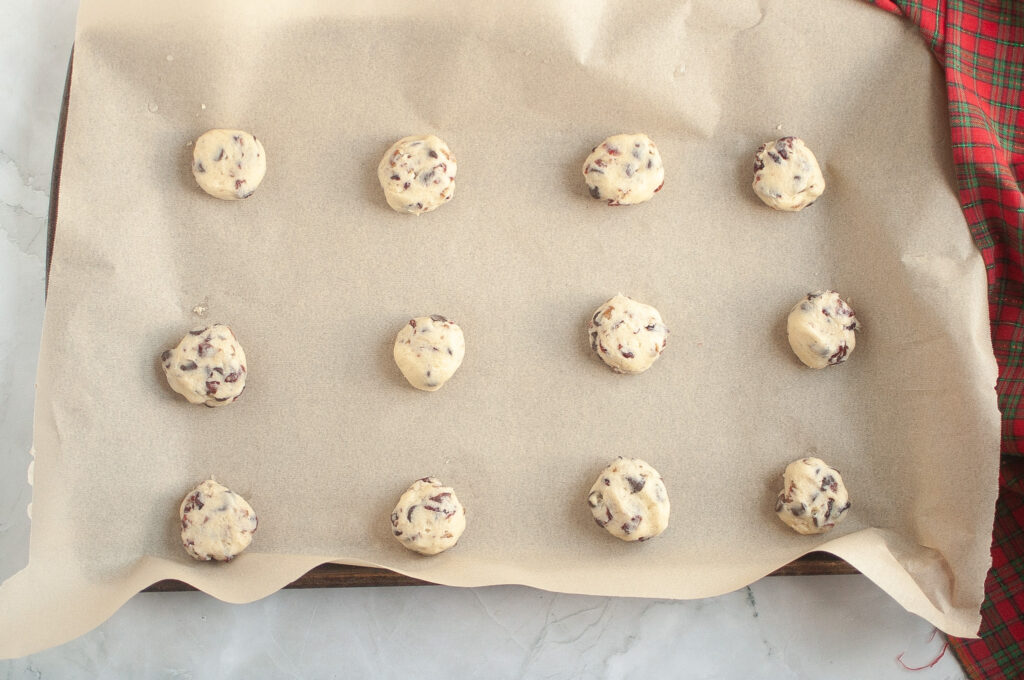Twelve unbaked chocolate chip cookie dough balls are arranged on a parchment-lined baking sheet, with a red plaid cloth in the corner.