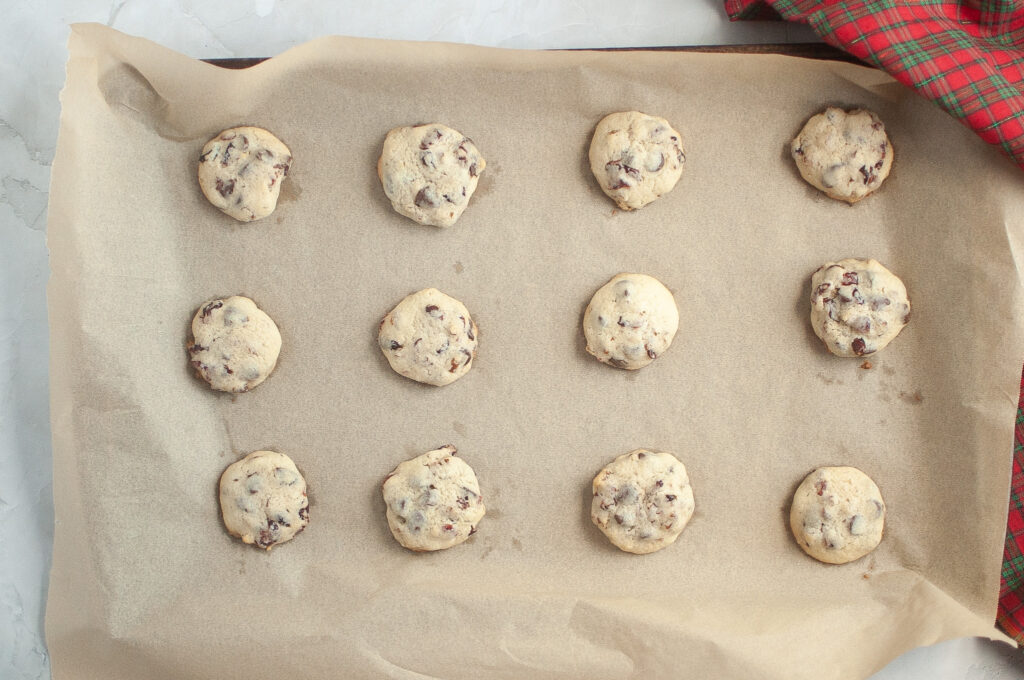 A baking tray lined with parchment paper holds twelve chocolate chip cookies arranged in rows, with a red and green cloth visible in the top right corner.